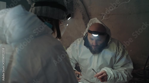 Caucasian researcher in protective suit contemplates contaminated vial while seated in ruined shelter under dim headlamp, bearded colleague watching, breath misting in cold, somber reflective mood