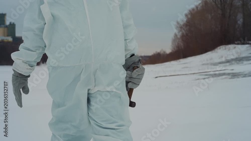 Scientist in protective suit holds metal cross near frozen field, closeup of gloved hand gripping rusted artifact, bleak snowplain, muted sky, abandoned perimeter suggesting nuclear fallout and tense