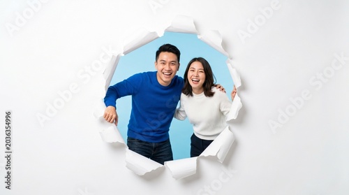 A Cheerful Couple Emerging Through Torn Paper: A happy Asian couple, a man and a woman, break through a torn paper opening, revealing their joyful expressions and vibrant blue background.