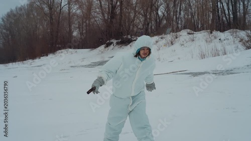 Person in white snowsuit walking, lone explorer strides across wide snowplain with small instrument in hand, distant trees and abandoned industrial silhouette on horizon, subtle sunset light