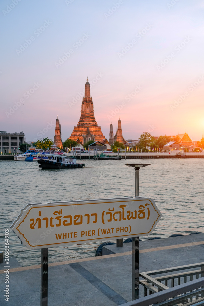 Obraz premium Wat Arun temple glows at dusk across the Chao Phraya River. A boat moves on the water near the White Pier sign, offering a scenic view of Bangkok, Thailand