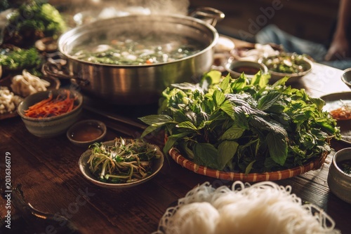 Family feast with communal pho broth pot as centerpiece and fresh herb basket with rice noodles arranged, Vietnamese family gathering tradition