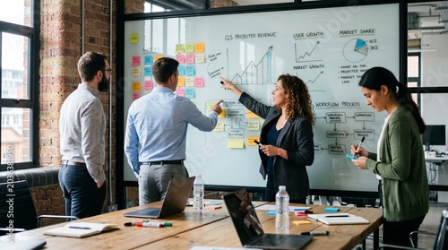 In the foreground, a large wooden conference table holds laptops, notebooks, markers, and water bottles, capturing a collaborative and dynamic brainstorming session.