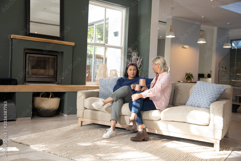 Fototapeta premium Mature female friends sipping coffee while chatting on sofa in modern living room with mugs