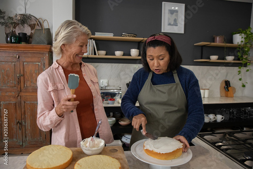Two mature bakers wearing navy and pink shirts frosting cake on kitchen counter, holding spatula