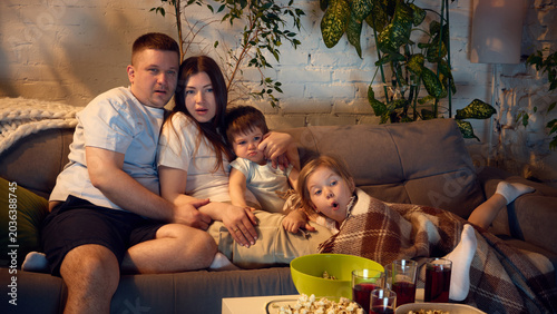 Fotografie Family watching movie at home with popcorn and red drinks on cozy sofa