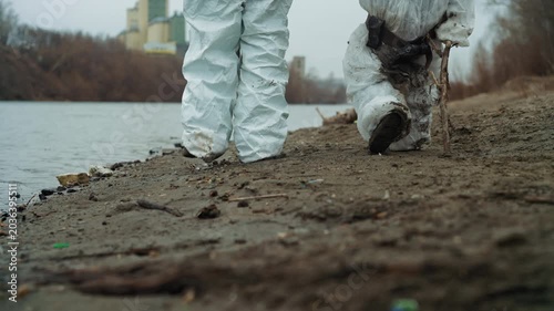 White scientist father and child riverside, wearing soiled protective suits, trudging through mud and debris, leaving heavy footprints beside polluted water, industrial plant looming in distance, cold