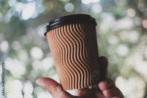 A single hand holding a brown paper cup with blurry green foliage background