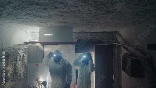 Two researchers in protective suits documenting contamination and collecting labeled vials, packing gear under dim headlamp light, recording data and preparing exit through ruined doorway with heavy
