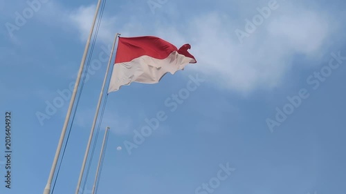 The Indonesian flag waves proudly against a bright blue sky, creating a peaceful and patriotic scene with soft clouds in the background.