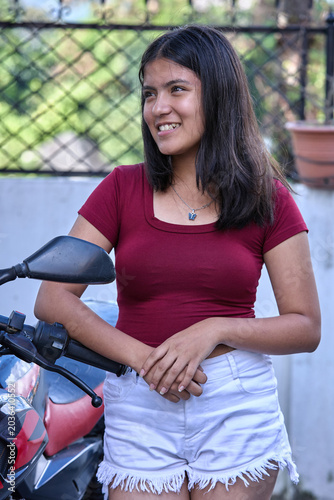 Young woman in red top and white shorts standing beside motorcycle outdoors hands clasped