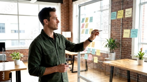 A man in a modern office writes on a transparent whiteboard covered with colorful sticky notes, planning or brainstorming ideas.