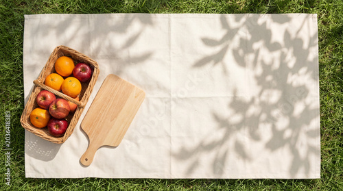 Flat lay top view of a white picnic blanket on green grass with a fruit basket, wooden board, and leaf shadows