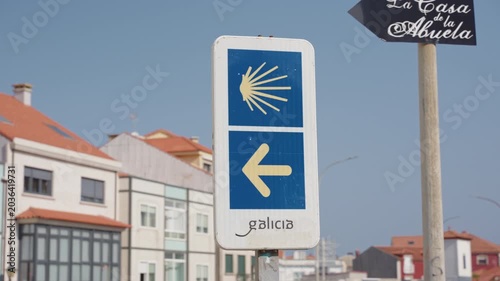 Camino de Santiago waymarker sign featuring a yellow scallop shell and left arrow on a blue background, located in a town in Galicia.