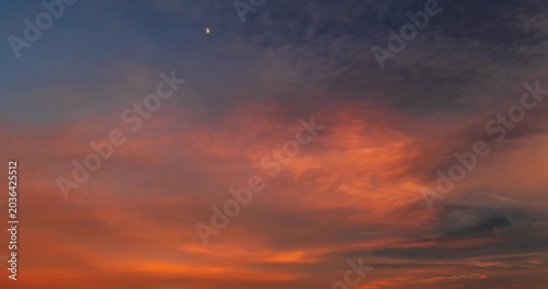 Scenic dramatic sky at twilight with vibrant red and purple sunset clouds and tiny moon high in the center.