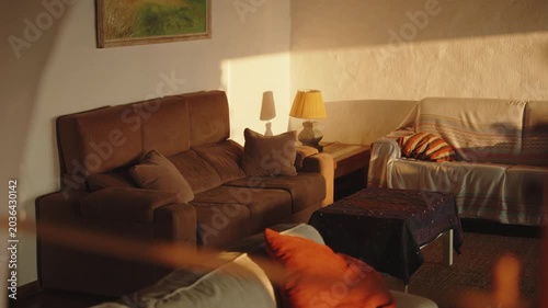 Warm, sunlit interior of a cozy living room at golden hour, featuring comfortable brown sofas and a lamp casting a soft glow, creating an atmosphere of peaceful relaxation and domestic tranquility.