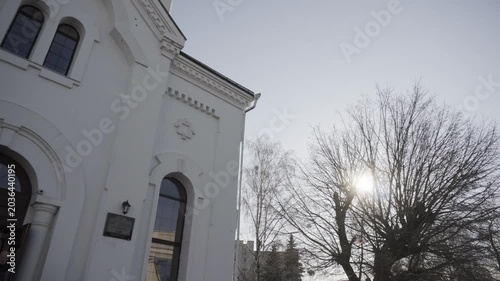 White orthodox church exterior. Golden onion dome, prominent bell tower, arched windows visible. Winter sunlight filters through bare tree branches. Serene, spiritual atmosphere for religious architec