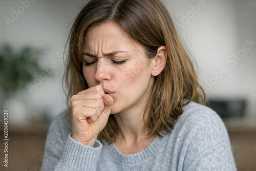 A woman sneezing into her hand, with red-nosed discomfort due to seasonal allergies or cold symptoms. She is wearing a cozy sweater and appears indoors in a warm setting.