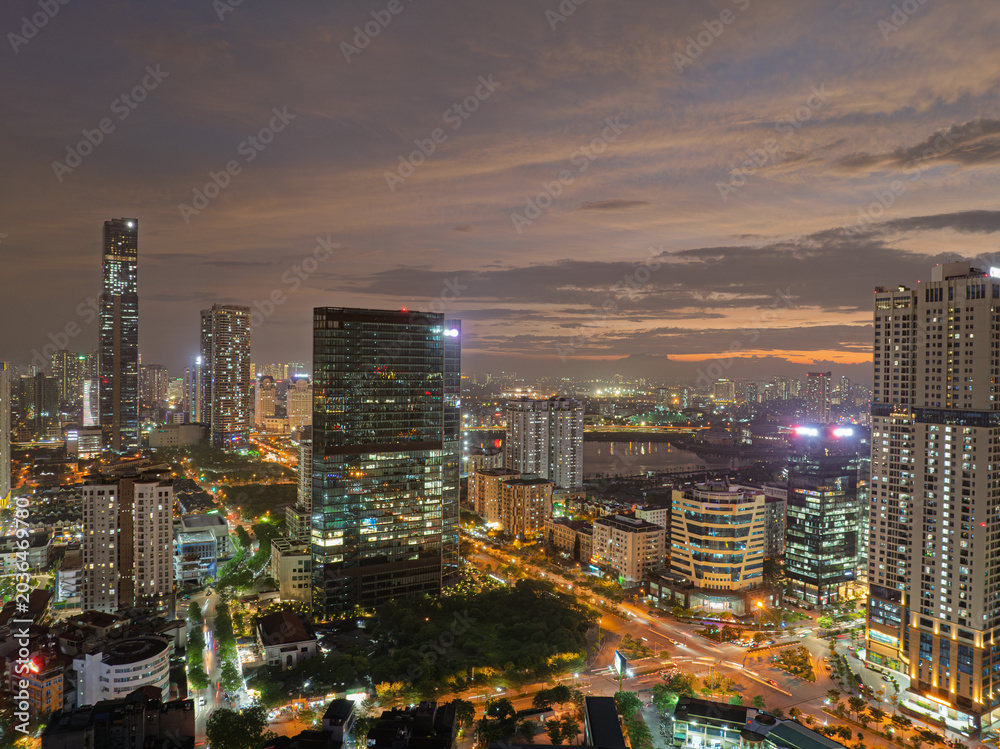 Obraz premium Wide aerial panorama of modern Hanoi skyline featuring multilane roads, residential towers, and green urban landscapes beside a lake.