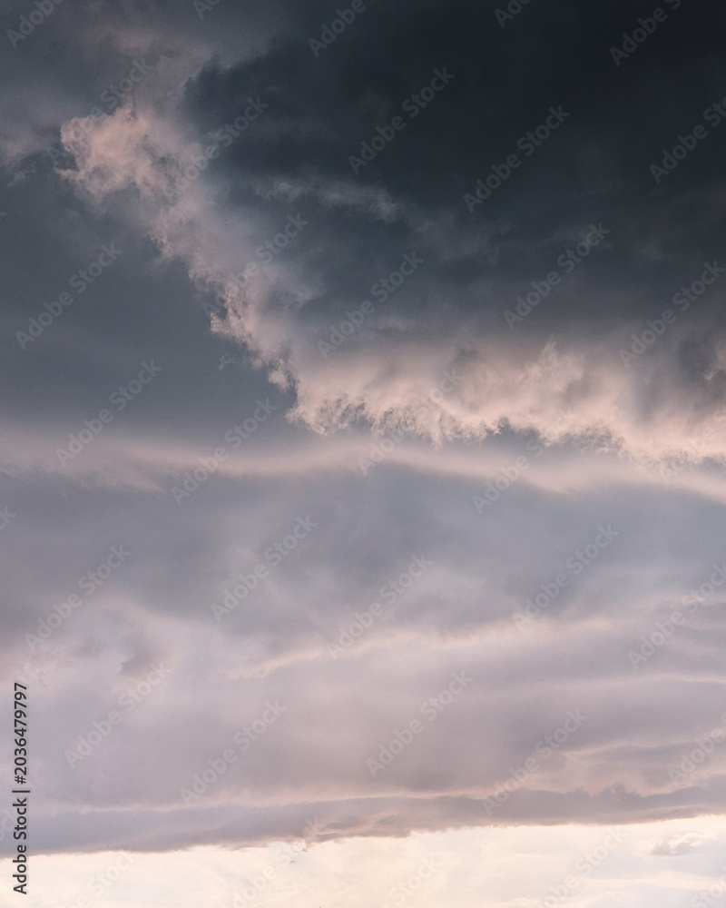 Obraz premium Moody storm clouds at sunset with soft pink light. Heavy, dark storm clouds rolling over a pale pink and grey sunset sky. Atmospheric, textured cloudscape before a summer rainstorm.