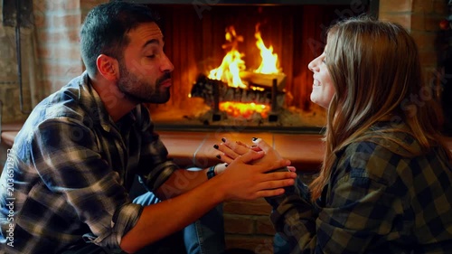 Romantic couple kissing in front of fireplace. Young happy couple relaxing and kissing by a cozy fireplace on a winter evening at home