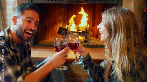 Romantic couple drinking wine and toasting by the fireplace. Happy young couple enjoying a romantic evening, toasting with glasses of red wine by a cozy fireplace