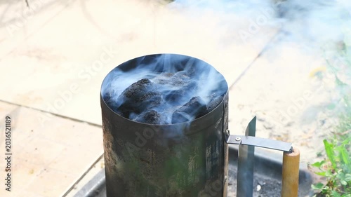 Charcoal briquettes glowing in a metal chimney starter, sending blue smoke skyward as the grill heats up for backyard bbqs, summer cookouts and weekend gatherings