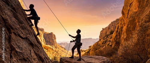 Two climbers scaling a rugged cliff face at sunset with safety ropes and harnesses on a rocky mountain