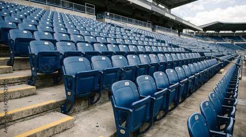 Rows of blue football stadium seats sit empty and quiet. Concrete steps lead to higher seating levels. Weathered metal and plastic show signs of use. No people fill the vast, open space