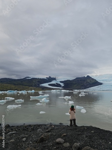 Woman traveler looking at floating icebergs in a glacial lagoon with a massive glacier and mountains in the background. Moody Nordic nature exploration and travel concept.