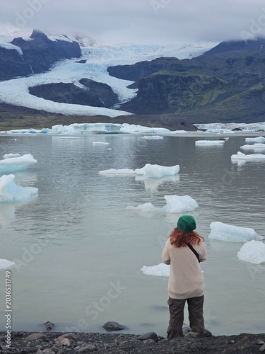 Woman traveler looking at floating icebergs in a glacial lagoon with a massive glacier and mountains in the background. Moody Nordic nature exploration and travel concept.