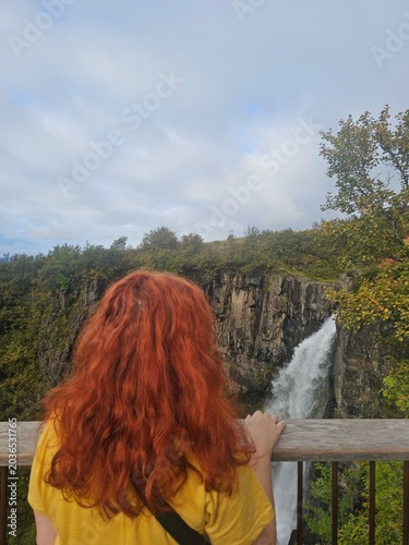 Red-haired woman traveler looking at a powerful waterfall from a viewpoint. Scenic Icelandic canyon landscape with rocky cliffs, autumn nature exploration and travel concept.