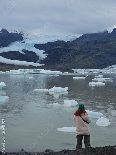 Woman traveler looking at floating icebergs in a glacial lagoon with a massive glacier and mountains in the background. Moody Nordic nature exploration and travel concept.