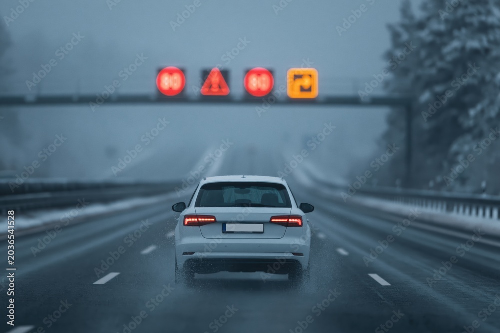 Fototapeta premium White car driving on foggy winter highway with warning signs and taillights