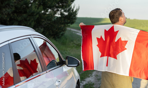 man stands near white car, holding a large Canadian flag, and their beautiful reflection in the car window. Happy Canada Day. Freedom, joy, national pride. Symbol of the country.