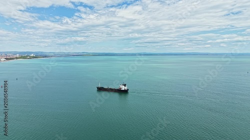 A cargo ship sails through clear blue water with small waves in the background. The sky is partly cloudy with a slight breeze. The coast is visible in the distance.