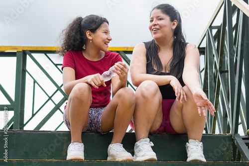 Obraz na płótnie Latina mother and daughter sitting on bridge stairs laughing together during cas