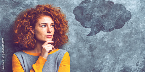 Thinking redhead woman looking at empty thought bubble on gray wall