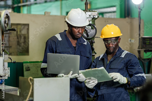 Foto Two African technicians stand with laptop and clipboard near industrial machine, checking document detail and digital record for maintenance review, production tracking, and factory coordination