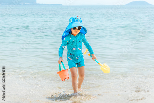 Child wearing blue swimwear and a sun hat walks through shallow water at the beach, holding a bucket and shovel, with a clear sky and distant hills visible in the background