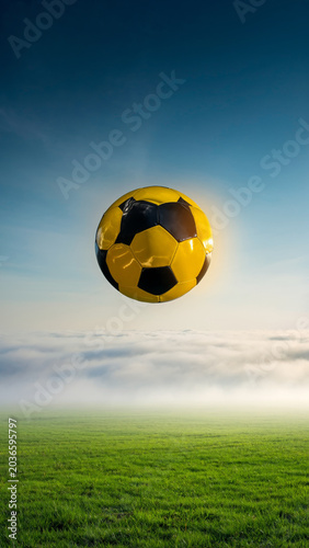 A vibrant yellow and black soccer ball hangs suspended in the bright blue sky above a misty green field, suggesting an aerial dynamic.