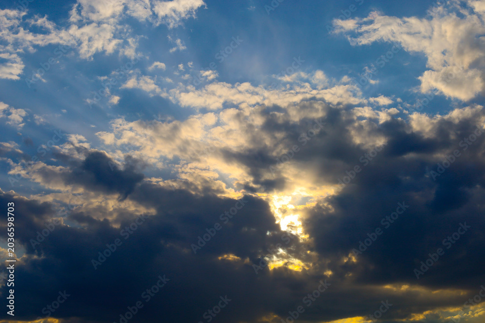 Obraz premium Bright sunbeams break through thick dark clouds in the evening sky. Contrasting golden light illuminates the edges of cumulus clouds, creating a dramatic effect.