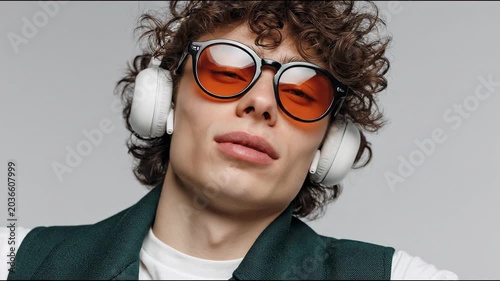 Young male model with curly hair wearing oversized sunglasses and headphones poses confidently against a neutral background, showcasing a stylish and modern look