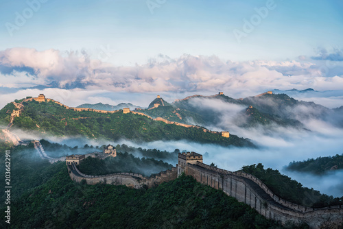 A breathtaking view of the Great Wall of China at Jinshanling during sunrise, with a sea of clouds engulfing the lush green hills and ancient fortifications.
