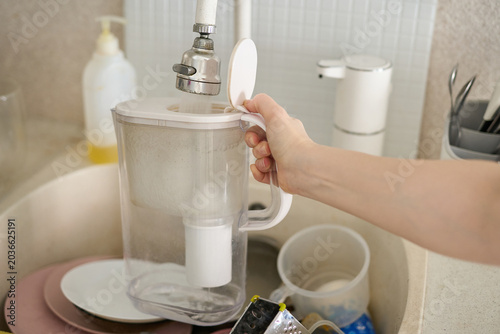 Water Filter Pitcher Being Held Open Under Faucet Over Kitchen Sink With Dishes Soap Dispenser And Visible Cartridge In Transparent Plastic Jug.