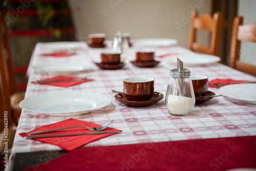 Cozy dining table prepared for breakfast or brunch with coffee cups, white plates and red tablecloth in a warm rustic interior. Inviting restaurant or family dining setting with wooden chairs, natural