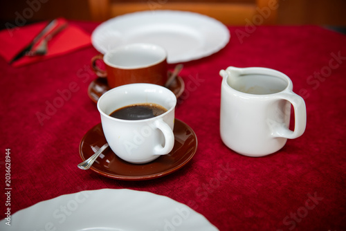 Cozy dining table prepared for breakfast or brunch with coffee cups, white plates and red tablecloth in a warm rustic interior. Inviting restaurant or family dining setting with wooden chairs, natural
