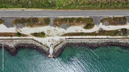 Aerial coastal highway turquoise sea shimmering sunlight glints on water, narrow road hugs rocky seawall, scattered vegetation and sandy patches, occasional