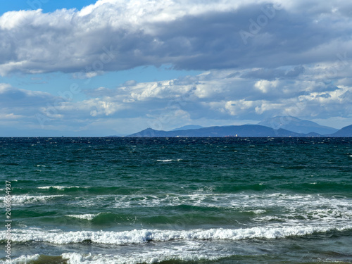 Canvas Print Choppy waves roll toward the shore under scattered clouds and blue sky near Voul