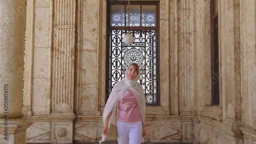Slow motion shot of female traveler walking toward camera inside historic mosque corridor in Cairo, Egypt. Elegant Islamic architecture and ornate marble details in cinematic 4K travel footage.